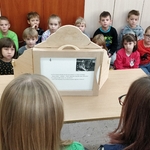 fWidownia z klasy I a patrzy na teatrzyk kamishibai Na pierwszym planie odwrocie teatrzyku i głowy czytających.jpg