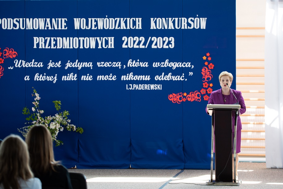 Kobieta przemawia - za nią biały napis na niebieskim tle.jpg