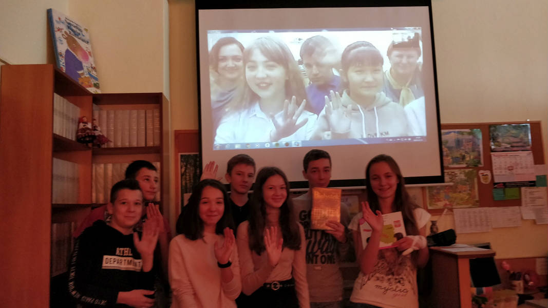 Uczniowie z książkami stoją pod monitorem, na którym widać dzieci z innych krajów łączące się na Skype.png