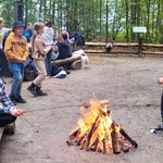 Nauczyciel i uczeń pieką kiełbaski nad ogniskiem w tle grupa dzieci przy stole.jpg