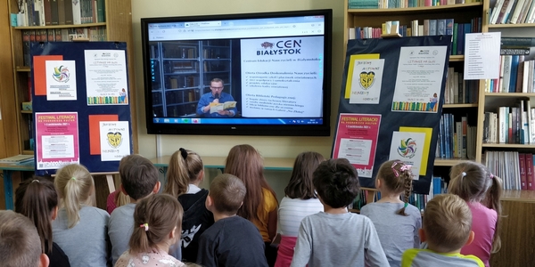 Dzieci patrzą na wielki monitor na którym aktor czyta książkę